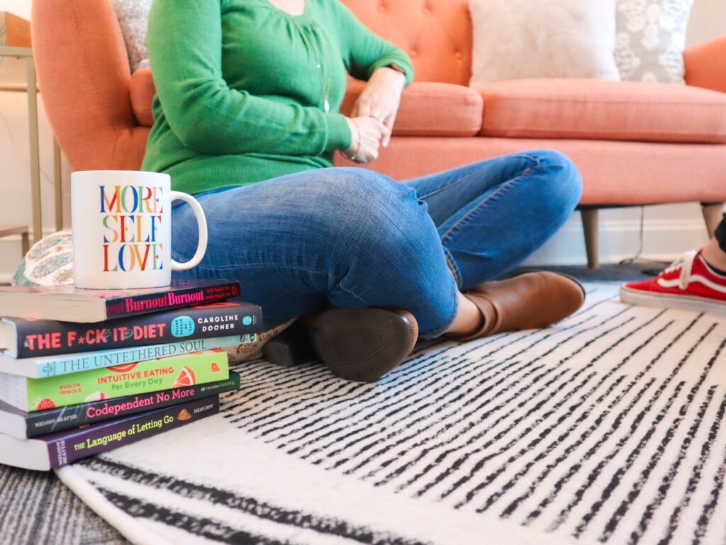 Woman sitting on a rug in a therapy room, stack of self-healing books and a coffee mug that says "more self love." Feeling your feelings blog from Evoke Wellness
